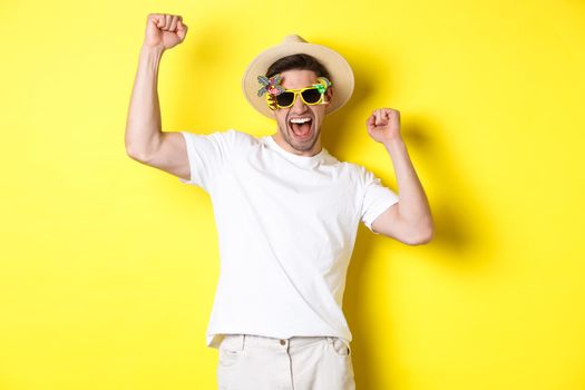 Concept of tourism and lifestyle. Happy lucky guy winning trip, rejoicing and wearing holiday outfit, summer hat and sunglasses, yellow background.