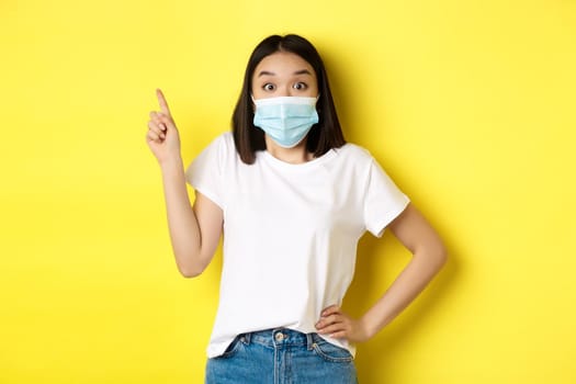 Covid, health care and pandemic concept. Asian female model in medical mask and white t-shirt pointing finger at upper left corner logo, showing promotion, yellow background.