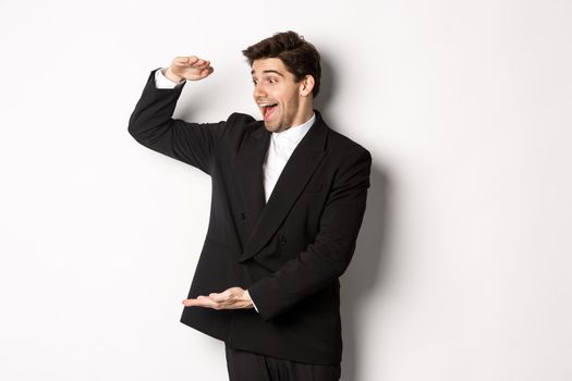 Portrait of excited handsome man in suit, shaping big object on copy space and smiling amazed, holding something, standing over white background.