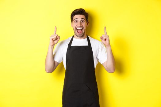 Excited coffee shop owner in black apron pointing fingers up, showing special offers, standing over yellow background.