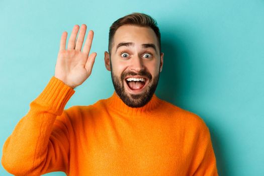 Handsome man waving hand to say hello, giving high five, standing over light blue background.