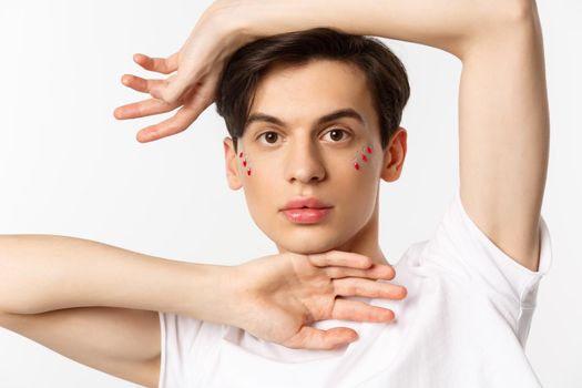 People, lgbtq and beauty concept. Close-up of beautiful androgynous male model with glitter on face, posing sensual at camera, standing over white background.