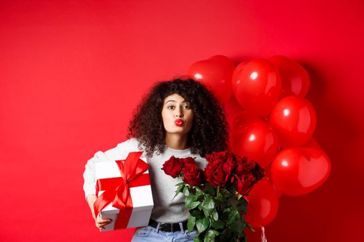 Holidays and celebration. Pretty woman celebrating birthday blowing air kiss, receive gifts and flowers on anniversary, standing near party balloons, red background.