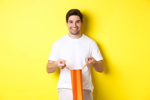 Happy young man open shopping bag with present, smiling at camera, standing against yellow background.