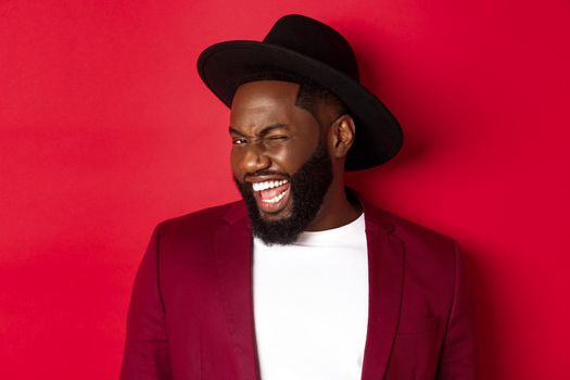 Cheeky african american man in party outfit, winking at camera and smiling, standing against red background.