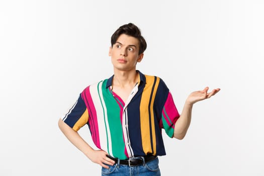 Confused gay man looking left at something strange, raising hand clueless and puzzled, standing over white background.