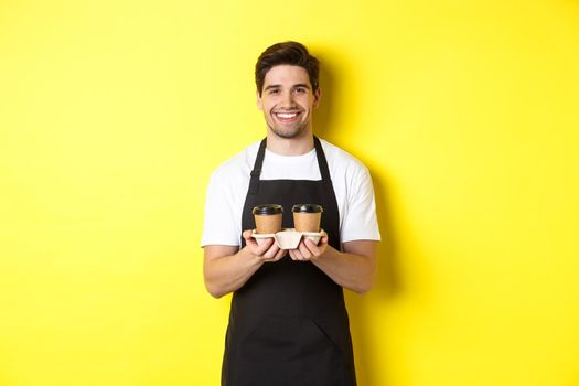 Handsome male barista serving takeaway coffee and smiling, bringing order, standing in black apron against yellow background.
