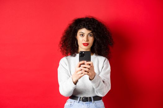 Young woman with curly hair, recording video on smartphone, taking photo on mobile phone and looking at camera, standing on red background.