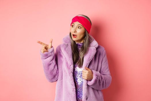 Fashion and shopping concept. Trendy elderly asian female in fake fur and glittering dress pointing, looking left with amazement, checking out offer, pink background.