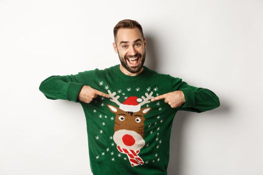 New year celebration and winter holidays concept. Cheerful man pointing at his funny Christmas sweater and smiling, white background.