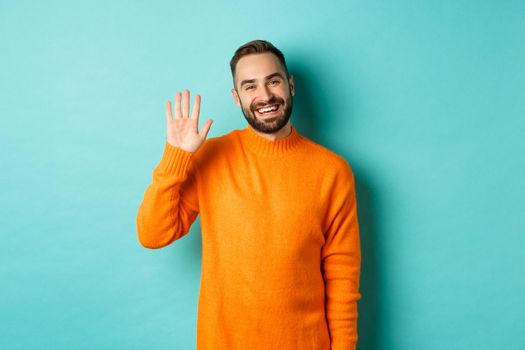 Photo of friendly young man saying hello, smiling and waiving hand, greeting you, standing in orange sweater over light blue background.