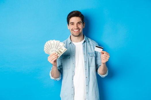Young smiling man showing cash dollars and credit card, standing over blue background.