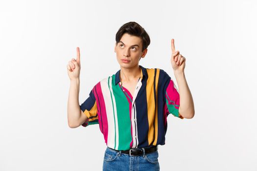 Attractive gay man looking thoughtful, pointing fingers up at logo, standing over white background. Copy space