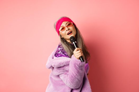 Stylish and sassy asian mature woman perform song on stage, holding mic and singing karaoke, wearing trendy heart-shaped sunglasses and faux fur coat, pink background.