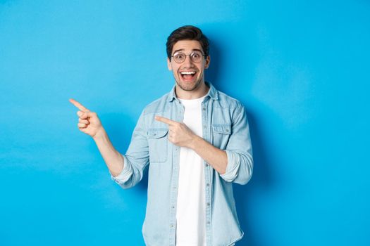 Excited handsome man in glasses pointing fingers left, looking happy while showing advertisement, demonstrating copy space on blue background.