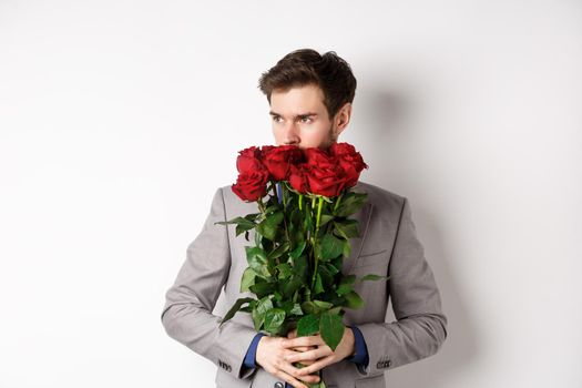 Romantic man in suit smell bouquet of roses and looking pensive, standing over white background. Concept of valentines day.