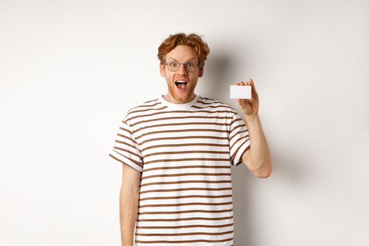 Shopping and finance concept. Amazed young man with beard and red hair showing plastic credit card and looking impressed, white background.