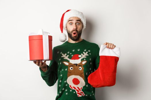 Xmas and winter holidays concept. Excited man holding christmas sock and gift box, celebrating New Year, standing over white background.