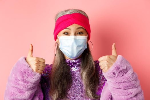 Covid-19, virus and fashion concept. Close-up of fashionable asian senior woman in face mask, showing thumbs-up and looking happy, pink background.