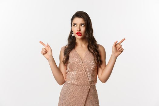 Christmas party. Thoughtful woman in glamour dress pointing fingers sideways, making choice, thinking and showing left and right offers, standing over white background.