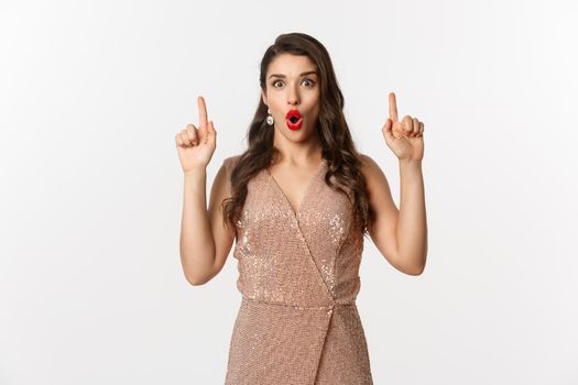 New Year, christmas and celebration concept. Surprised and excited woman in glamour dress, wearing outfit for party, pointing fingers up, showing promo offer, white background.
