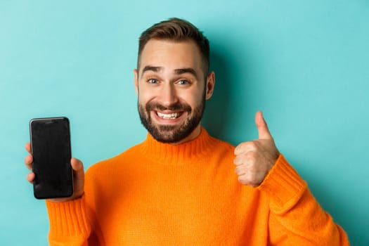 Close-up of attractive bearded man showing smartphone screen, thumbs up, recommending mobile app, standing satisfied over turquoise background.