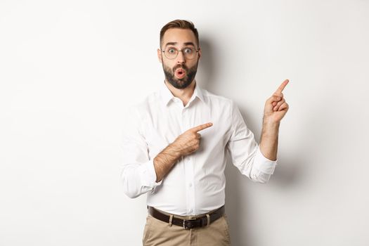 Impressed businessman showing interesting promo offer, pointing fingers right, standing amazed against white background.