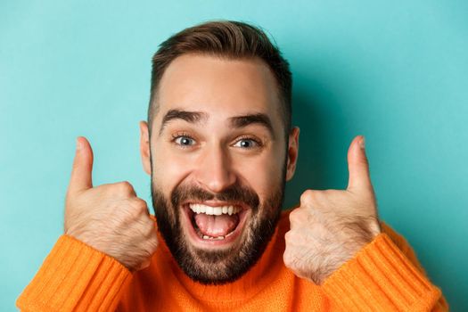 Headshot of excited bearded man showing thumbs-up, praising something good, approve and like product, standing over turquoise background.