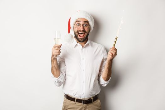 Winter holidays and celebration. Handsome bearded man having New Year party, holding firework sparkler and champagne, wearing santa hat, white background.