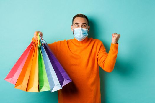 Covid-19, social distancing and lifestyle concept. Excited man in face mask showing shopping bags and rejoicing from discounts, standing over turquoise background.