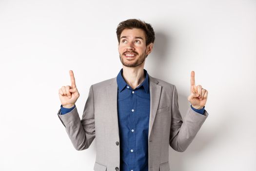 Dreamy smiling male manager in suit pointing and looking up, happy face, checking out advertisement, standing against white background.