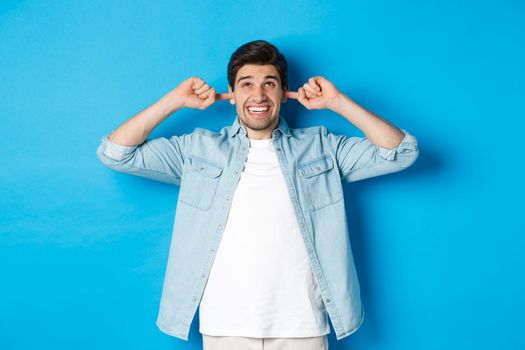 Irritated man close ears from noisy neighbours, looking up and grimacing, hearing loud music, standing over blue background.