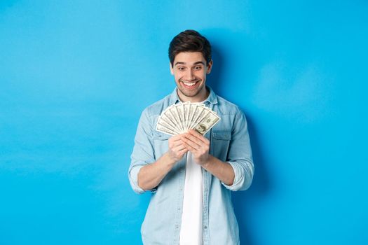 Excited successful man counting money, looking satisfied at cash and smiling, standing over blue background.