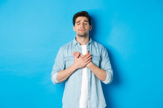 Portrait of dreamy and nostalgic guy in casual outfit, holding hands on heart and close eyes, daydreaming against blue background.