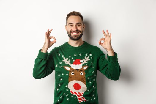 Christmas, holidays and celebration. Satisfied smiling man in green sweater showing OK signs and nodding in approval, recommending product, standing over white background.