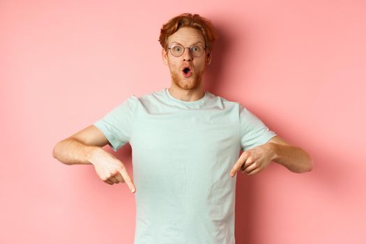 Image of amazed young man with red hair and beard, wearing glasses and t-shirt, pointing fingers down and staring excited at camera, pink background.