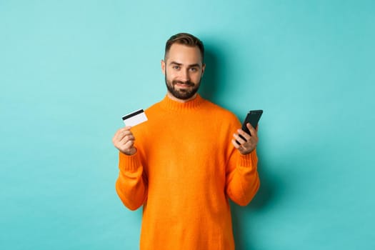 Online shopping. Handsome caucasian man in orange sweater, using credit card and mobile phone, standing over light blue background.