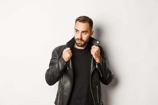 Attractive bearded man in leather biker jacket looking aside, standing confident against white background.