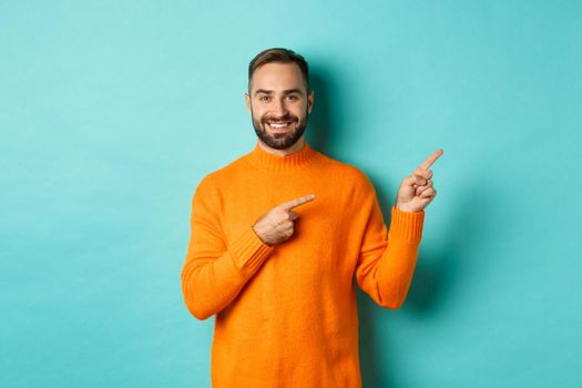 Confident happy guy in orange sweater, showing logo banner, pointing fingers right at copy space, turquoise background.