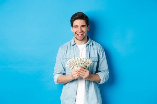 Happy successful man smiling pleased, holding money, standing over blue background.