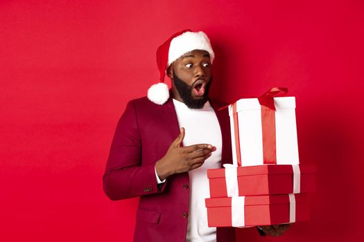 Christmas, New Year and shopping concept. Surprised Black man looking at xmas presents with amazement, wearing santa hat, standing with gifts against red background.