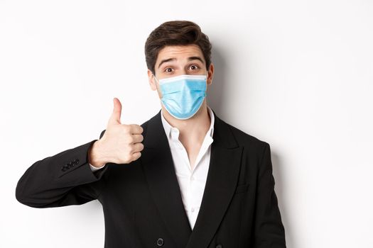 Concept of covid-19, business and social distancing. Close-up of happy businessman in black suit and medical mask, showing thumbs-up, making a compliment, white background.