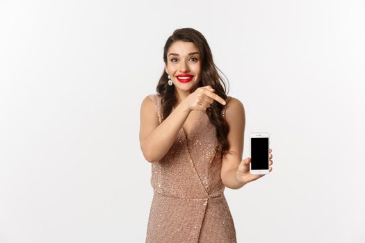 Online shopping. Excited young woman with red lips, elegant dress, pointing finger at mobile screen, showing something, standing over white background.