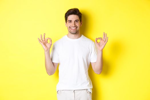 Relaxed guy smiling, showing okay signs, approve or agree, standing against yellow background.