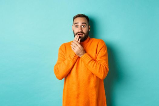 Scared and shocked guy looking at camera, standing in orange sweater against turquoise background.