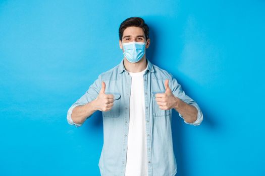 Concept of covid-19, pandemic and social distancing. Young man in medical mask showing thumbs-up in approval, like and agree, standing against blue background.