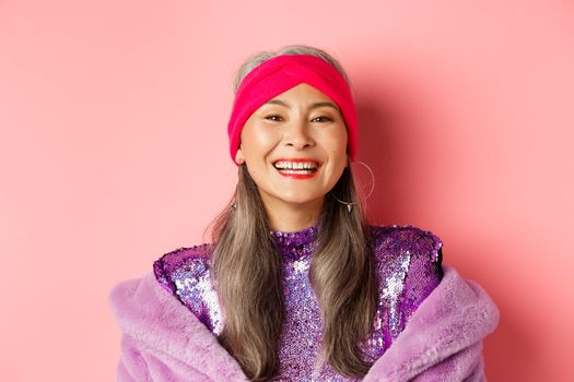 Close-up of happy asian senior lady in trendy purple outfit, laughing and smiling at camera, having fun on pink background.