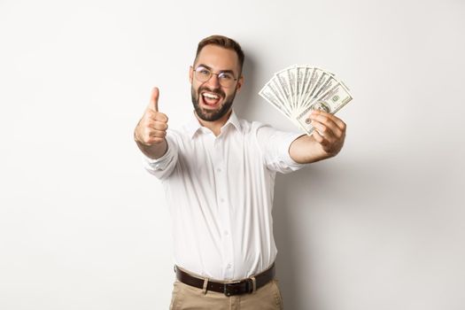 Excited man showing thumbs up and money, earning cash, standing over white background.