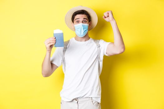 Concept of covid-19, travelling and quarantine. Happy man tourist in medical mask celebrating, showing passport with tickets for vacation and rejoicing, travel during coronavirus.