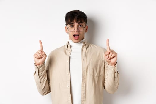 Amazed and excited man in glasses showing awesome promo deal, pointing fingers up and open mouth in awe, standing against white background.